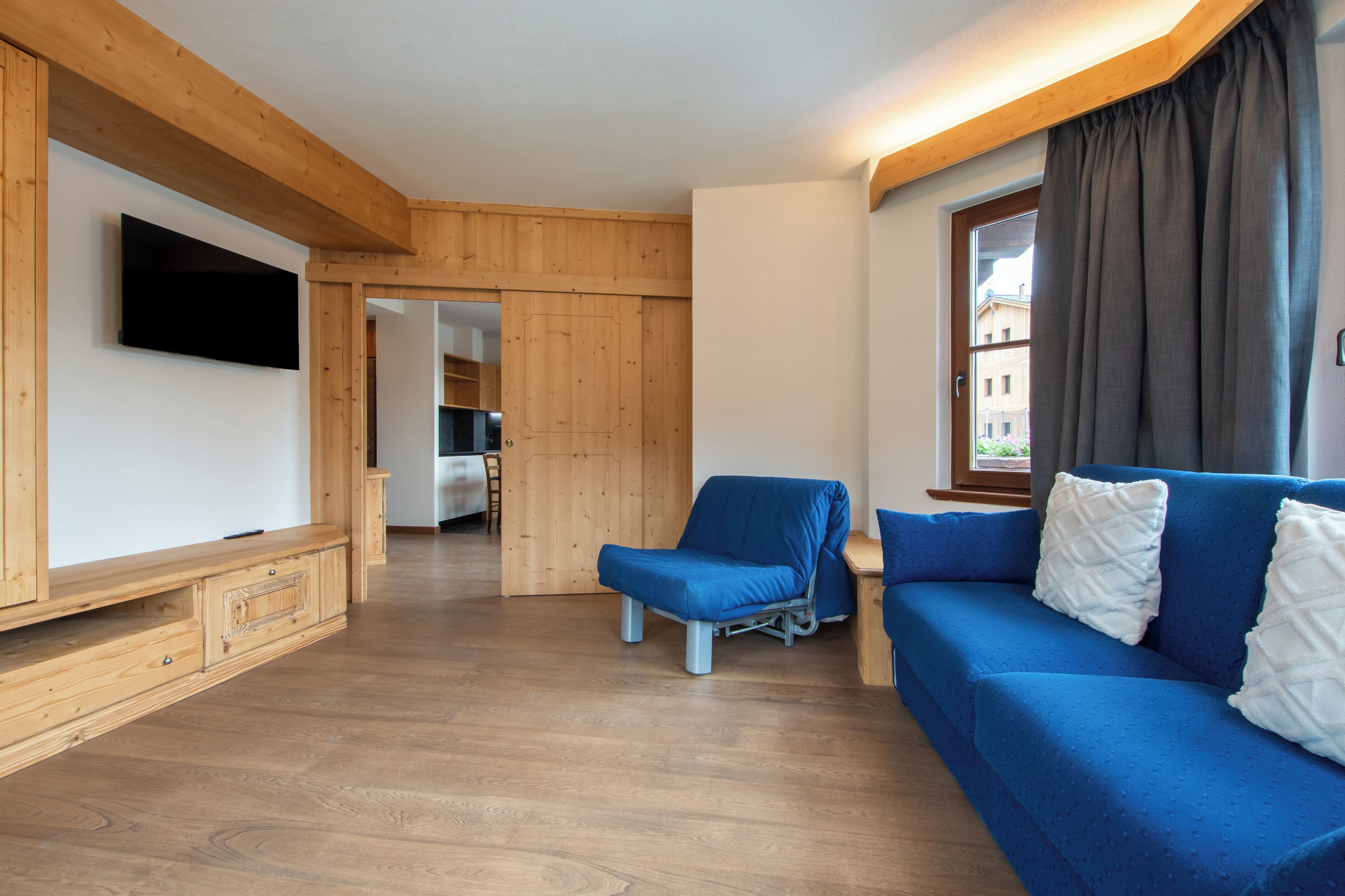 A room with a wooden floor, a blue sofa, a television, and a window with curtains.