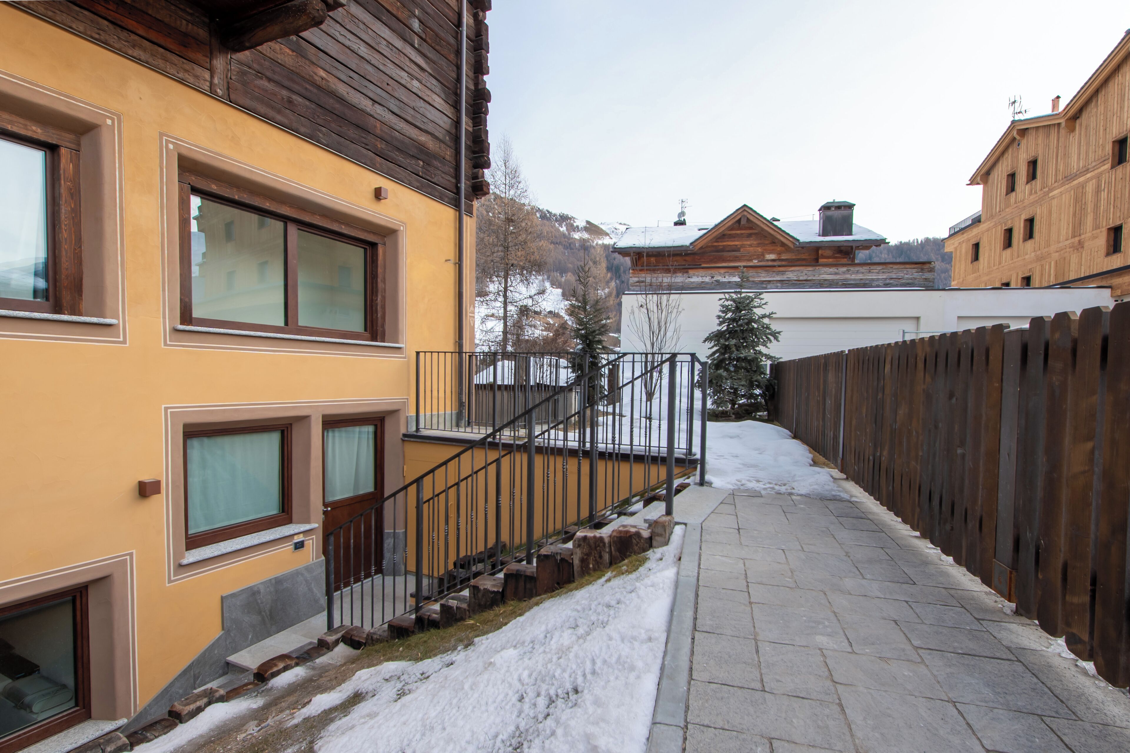 A snow-covered pathway leading to a yellow building with a wooden fence and a staircase.