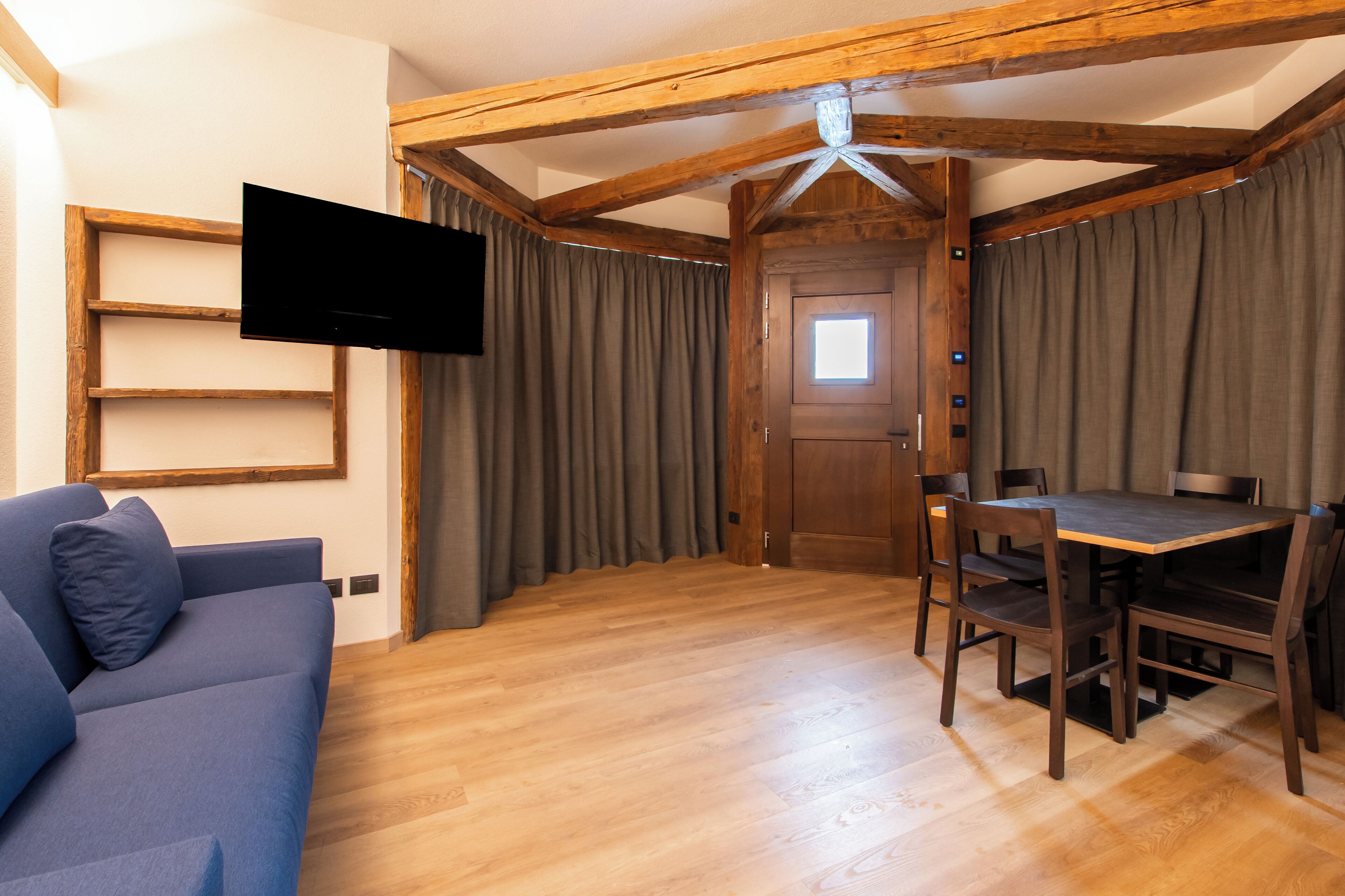 A room with a wooden ceiling, a flat-screen TV mounted on the wall, a blue sofa, a wooden dining table with chairs, and a door with a window.