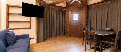 A room with a wooden ceiling, a flat-screen TV mounted on the wall, a blue sofa, a wooden dining table with chairs, and a door with a window.