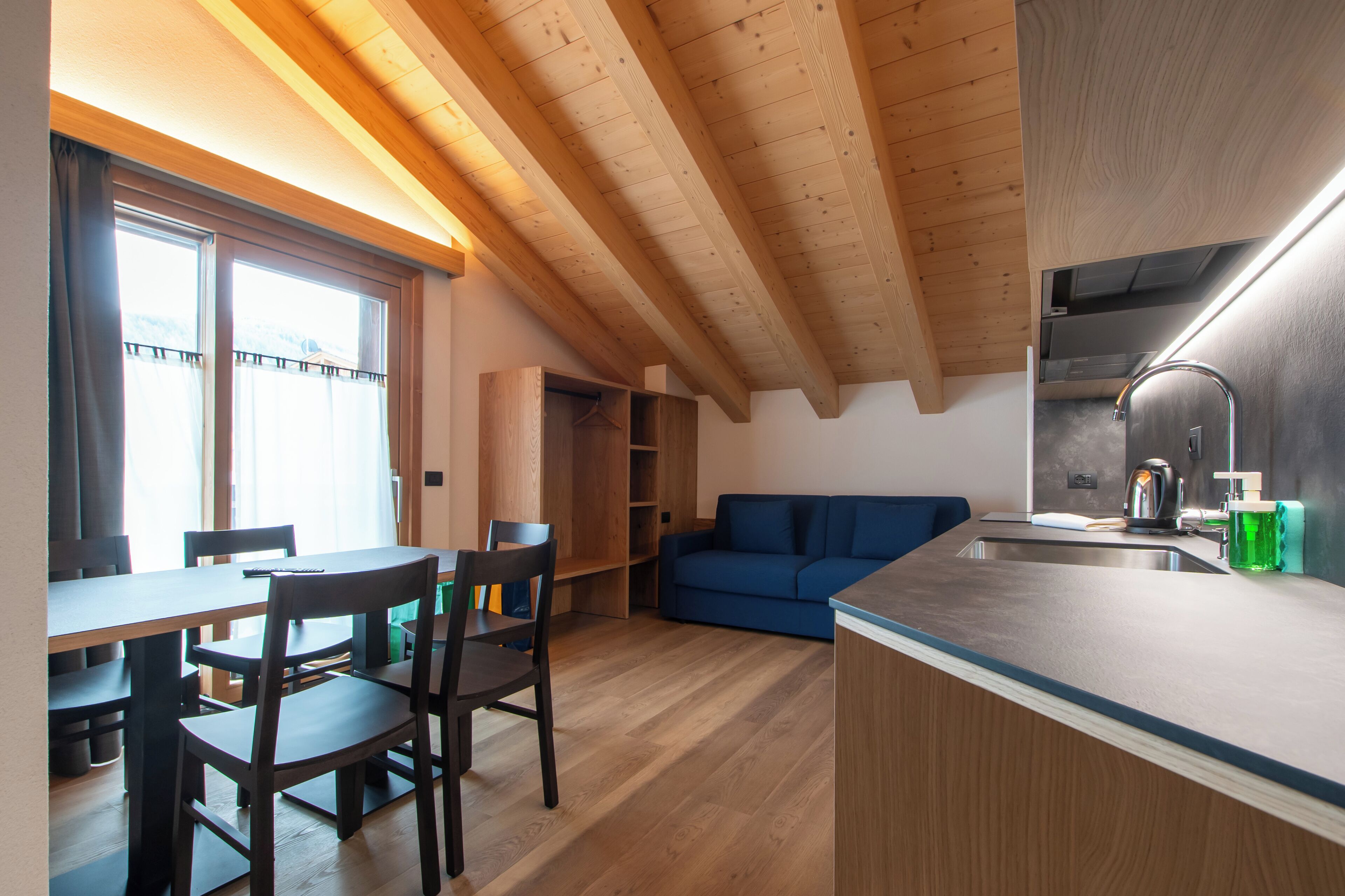A compact kitchen with wooden beams, a dining area with chairs, and a blue sofa.