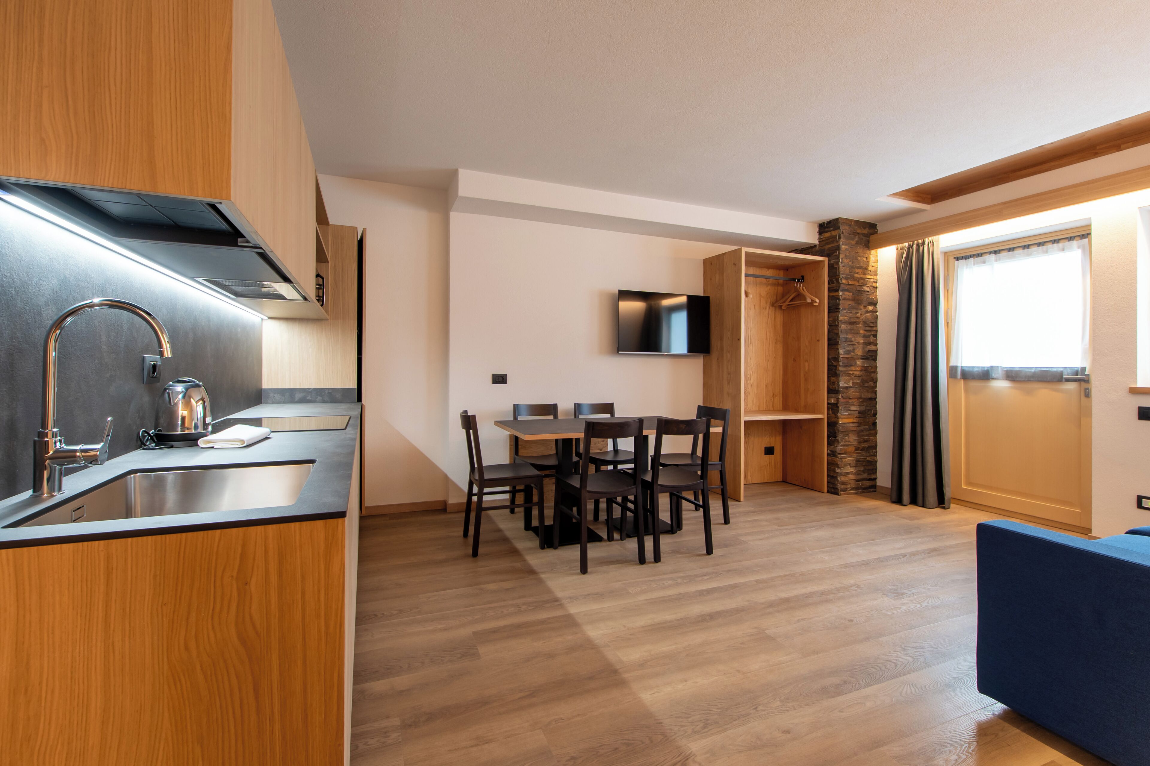 A modern kitchen with a built-in stove, a sink, and a coffee maker. There is a dining area with a table and chairs, and a TV mounted on the wall.