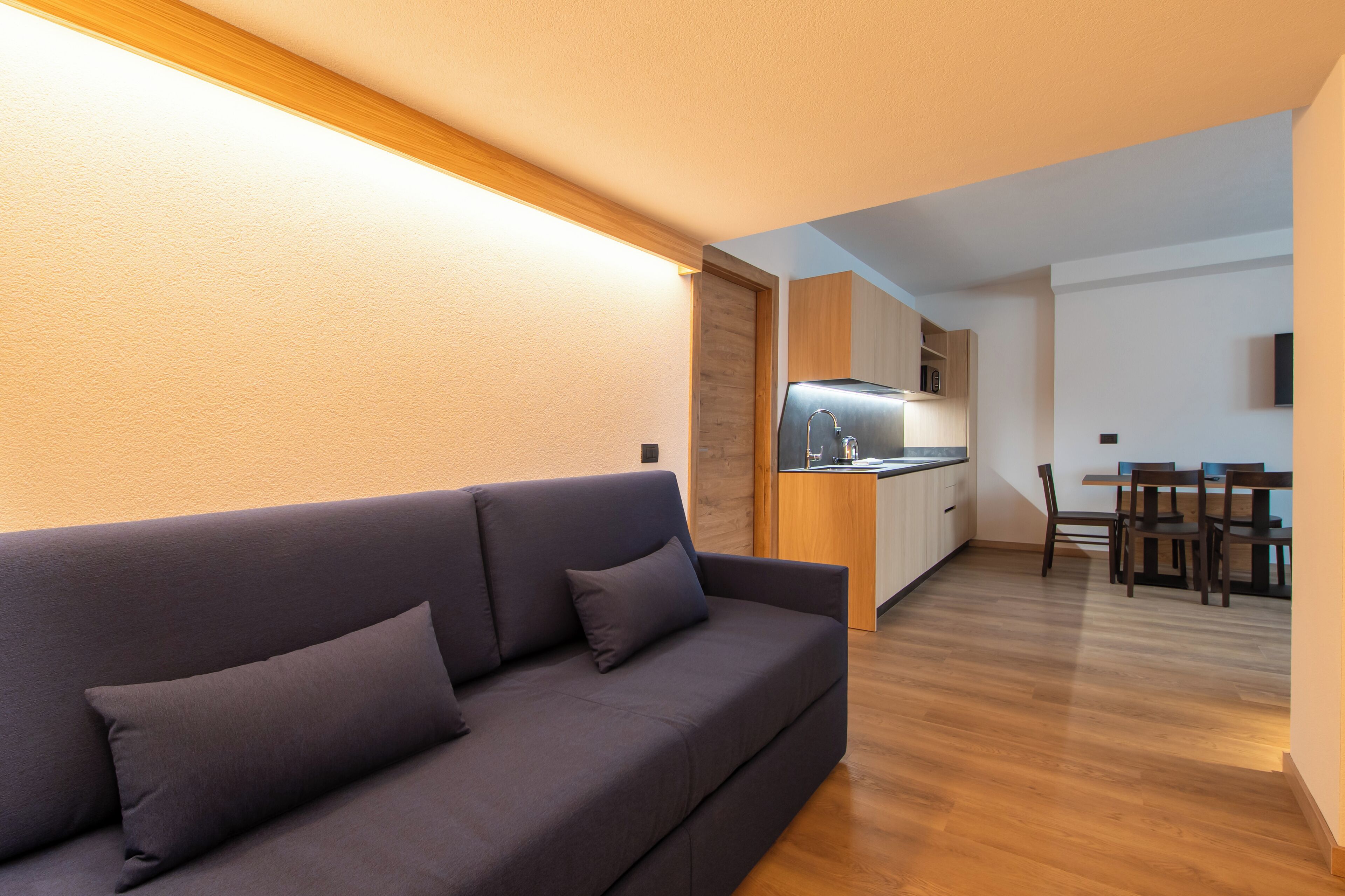 Ein modernes Wohnzimmer mit einem dunkelgrauen Sofa, Holzboden und einer Küche im Hintergrund.