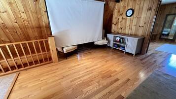 A room with wooden flooring, a white curtain, a clock on the wall, and a small cabinet with bottles.