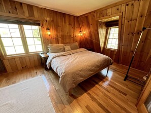 Une chambre avec des murs en bois, un lit, deux fenêtres et un radiateur portatif.
