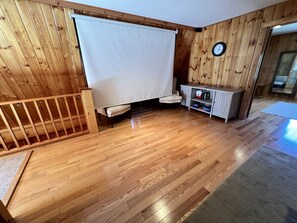 A room with wooden flooring, a white curtain, a clock on the wall, and a small cabinet with bottles.