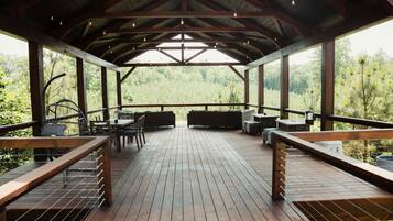 A covered outdoor deck with wooden flooring, seating arrangements, and large windows offering a view of the surrounding greenery.