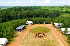 Un terrain de camping comprenant plusieurs cabanes, un espace central aménagé avec des chaises, et un sentier de terre qui les relie.