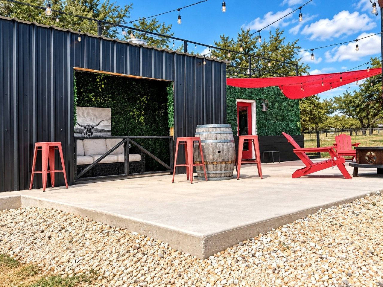 Une terrasse extérieure dotée d’un sol en béton, entourée de gravier, et comprenant un tonneau en métal, des chaises rouges et un auvent rouge.