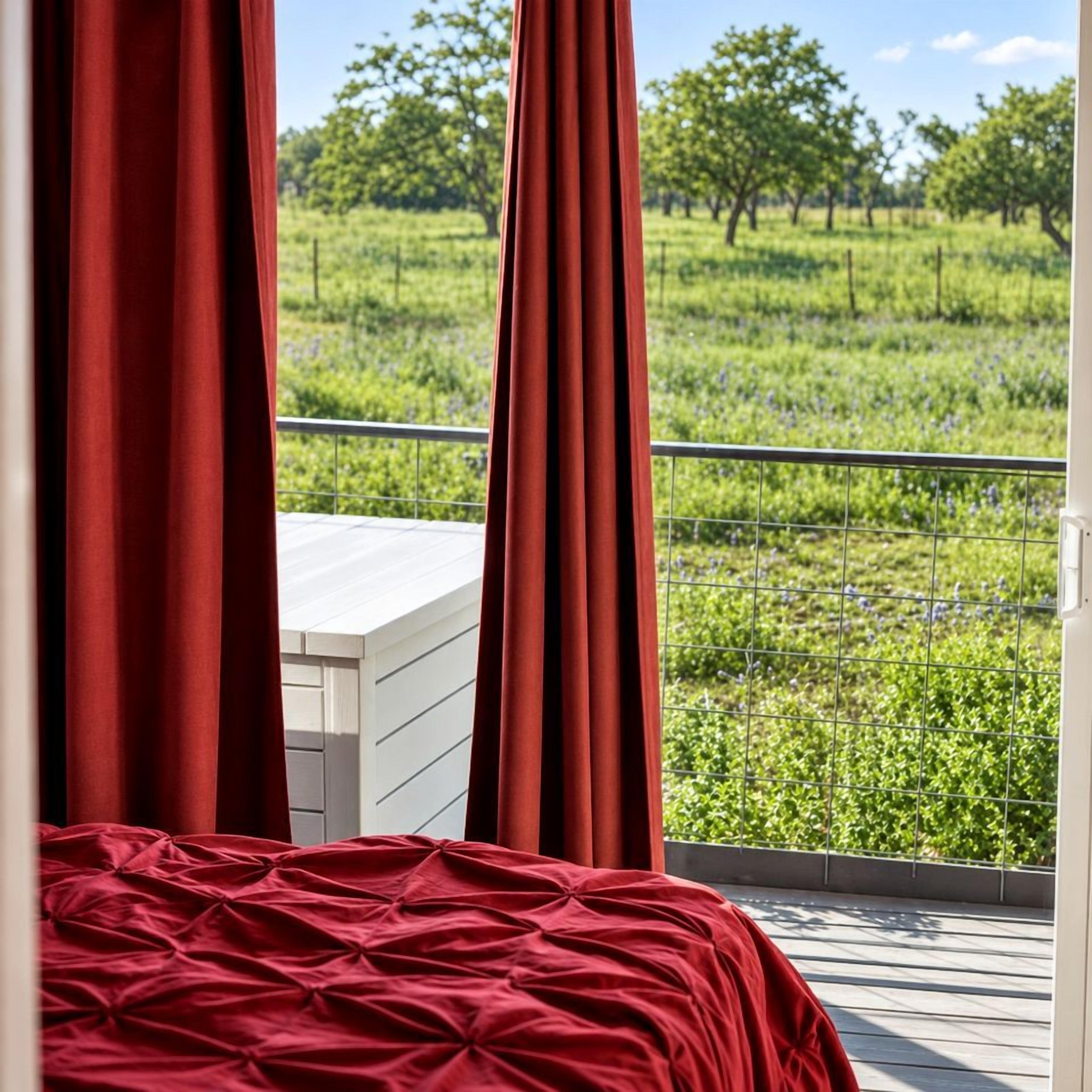 A bedroom with a bed, red curtains, and a view of a green field through a balcony railing.