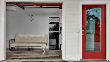 A beige sofa in a room with a red door, a kitchen area with appliances, and a wall-mounted clock.