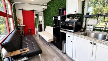 A compact kitchen with a black refrigerator, a coffee machine, and a sink. There is a white cabinet with a granite countertop. A black sofa and a white chair are placed in the living area.