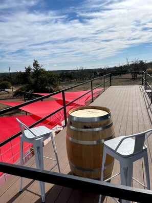 Una terraza de madera con barandillas metálicas, un barril de madera y sillas de plástico blancas.