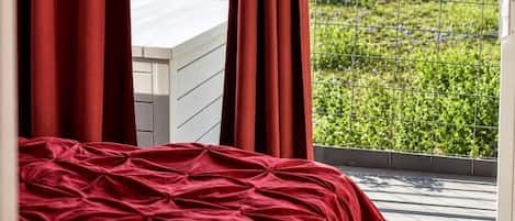 A bedroom with a bed, red curtains, and a view of a green field through a balcony railing.