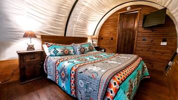 A tent-style room with a bed, wooden walls, a TV mounted on the wall, and a nightstand with a lamp.