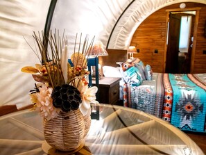 A tented accommodation with a bed, a table with a woven vase, and a lamp.