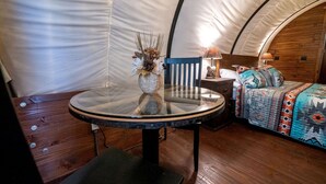 A tent with a round glass table, a chair, a bed with a patterned blanket, and a small lamp on a wooden nightstand. - Cozy Covered Wagon for a Unique Glamping Experience in Beaumont, Kansas (Beaumont)