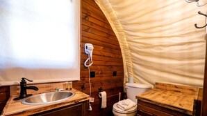 A compact bathroom with a wooden vanity, a sink, a toilet, and a hairdryer mounted on the wall. - Cozy Covered Wagon for a Unique Glamping Experience in Beaumont, Kansas (Beaumont)
