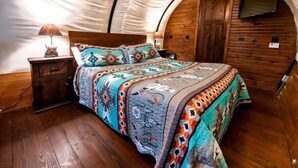 A tent-style room with a bed, wooden walls, a TV mounted on the wall, and a nightstand with a lamp. - Cozy Covered Wagon for a Unique Glamping Experience in Beaumont, Kansas (Beaumont)