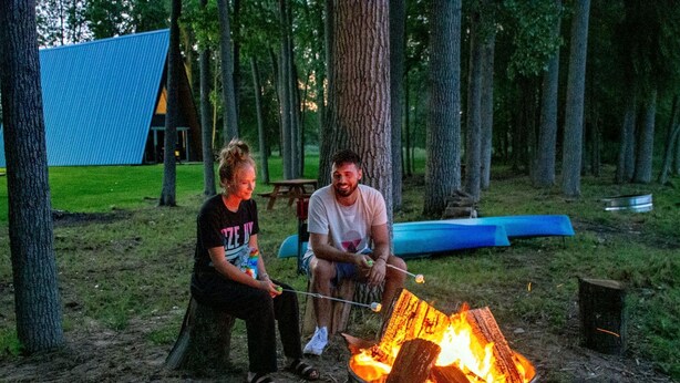 Two people roasting marshmallows over a campfire in a forested area with a tent and picnic table in the background.