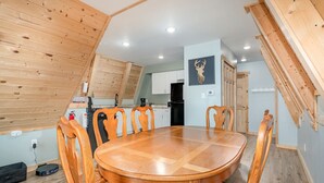 A dining area with a round wooden table and chairs, a kitchen in the background, and a ceiling with wooden beams.