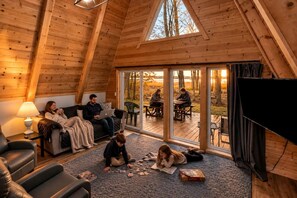 Ein gemütliches Blockhaus-Interieur mit Holzbalkendecke, einer Couch, einem Esstisch und einem großen Fenster mit Blick auf eine Terrasse.