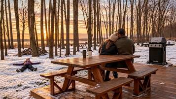 A wooden picnic table in a snowy outdoor setting with a grill and benches.