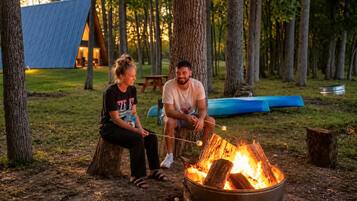 Zwei Personen sitzen an einem Lagerfeuer in einem bewaldeten Gebiet, im Hintergrund sind ein Zelt und ein Kanu zu sehen.