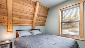 A bedroom with a sloped ceiling, a bed with grey bedding, a bedside table with a lamp, and a window with blinds.