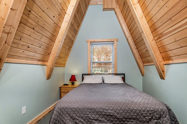 A bedroom with a sloped ceiling, a bed, a nightstand, a lamp, and a window.