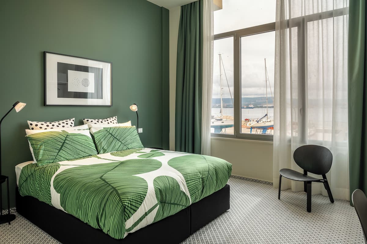 A modern bedroom with a green theme, featuring a large bed, a framed artwork, and a view of the harbor through a window.
