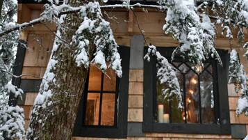 Una cabaña de madera con un ventanal grande y una puerta, rodeada de árboles cubiertos de nieve.