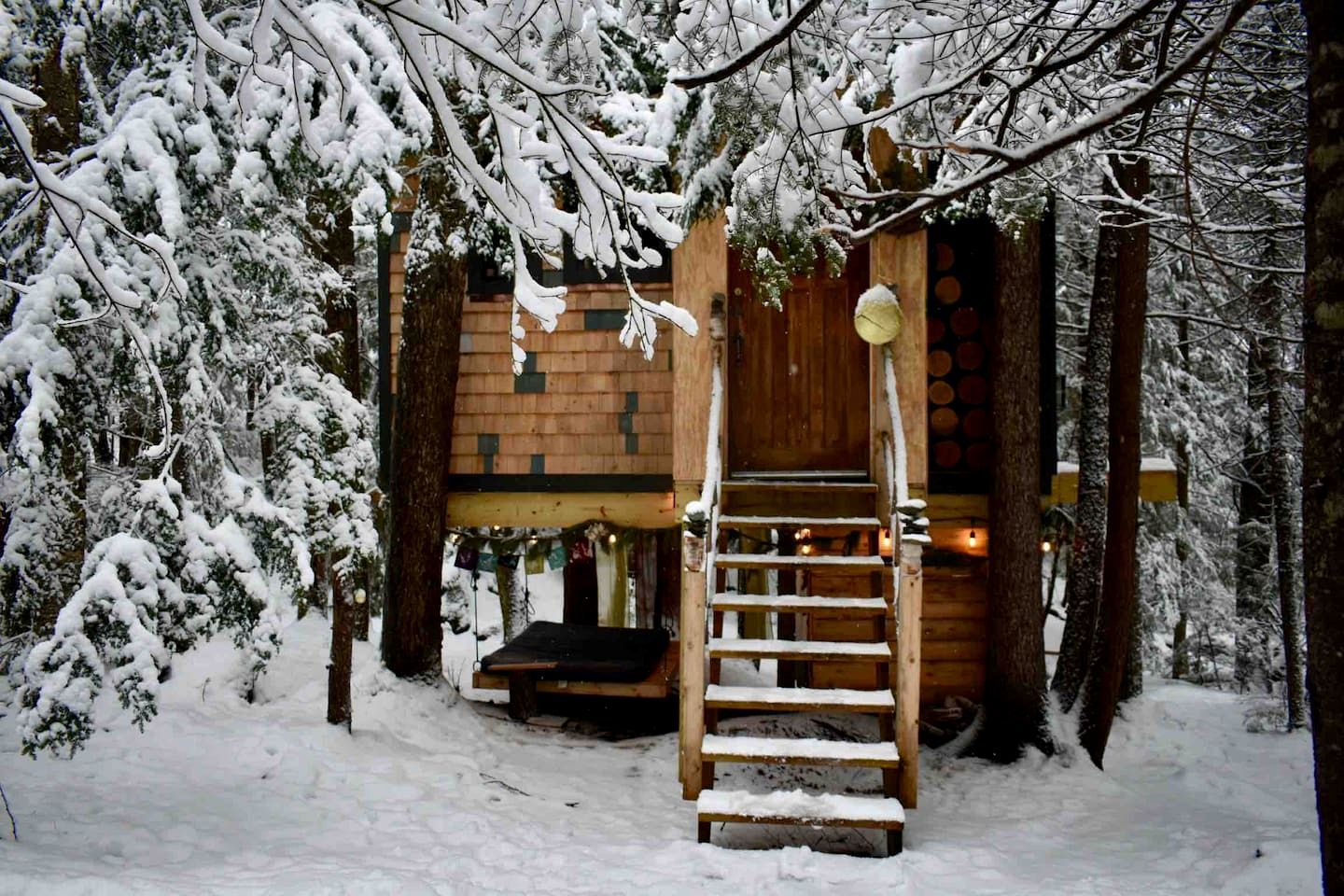 A wooden cabin with a wooden staircase leading to a door, surrounded by snow-covered trees.