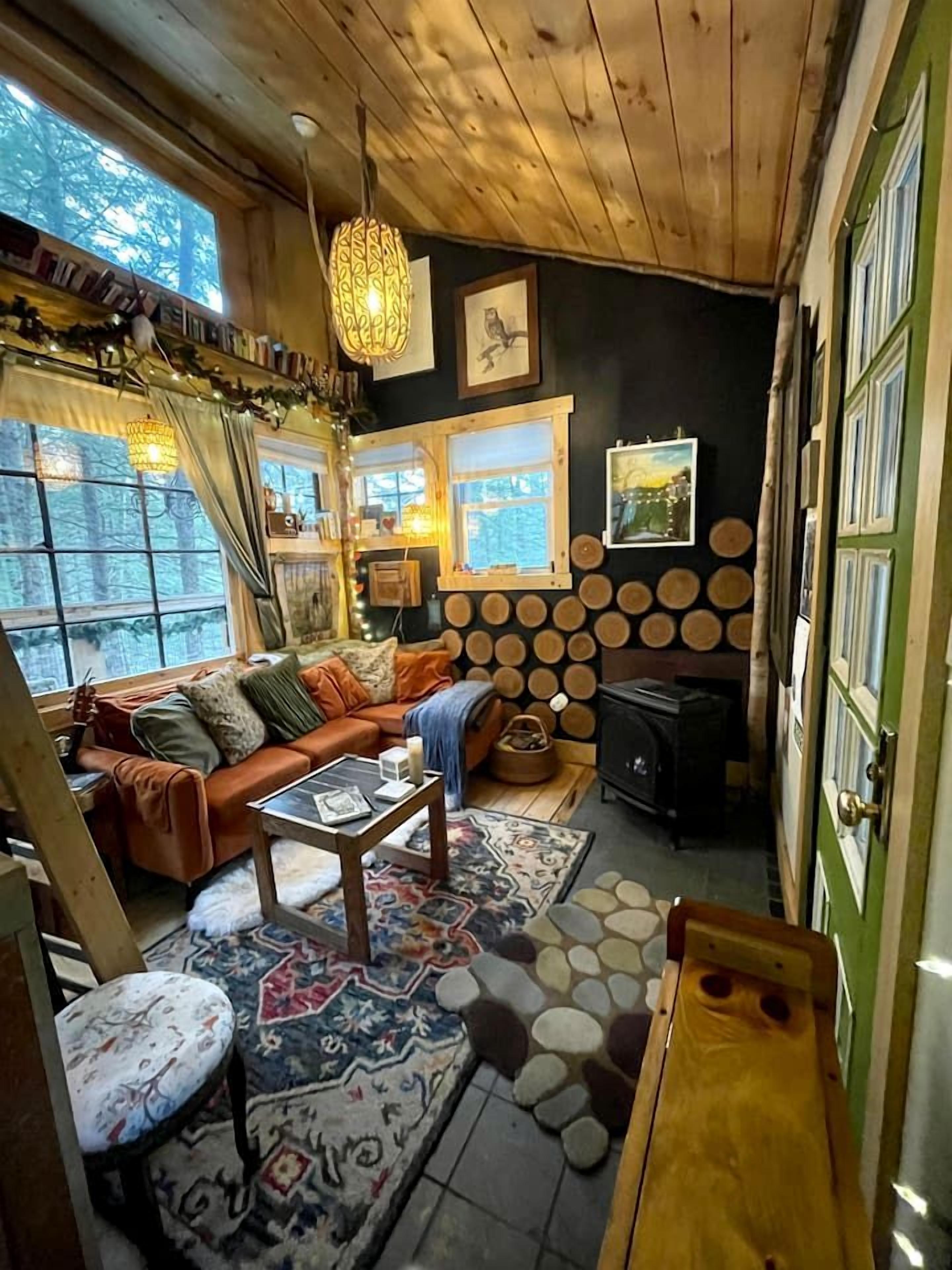 A cozy cabin interior with a wooden ceiling, a sofa, a coffee table, a fireplace, and a view of the forest through the windows.