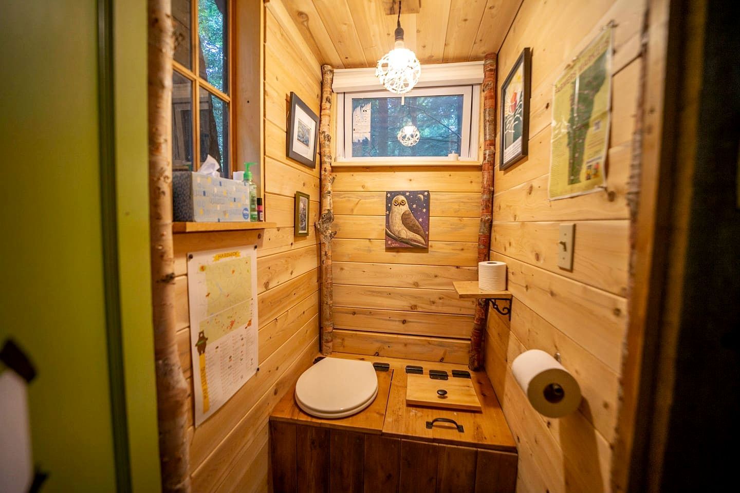 A small, wooden cabin-style bathroom with a toilet, a sink, a window, and a hanging light fixture.