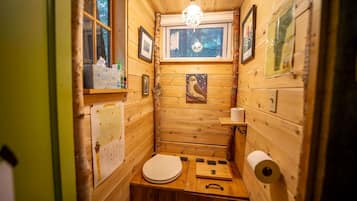 A small, wooden cabin-style bathroom with a toilet, a sink, a window, and a hanging light fixture.