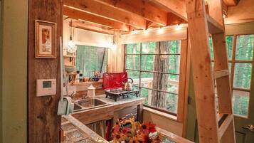 Interior rústico de una cabaña con zona de cocina, una mesa de comedor y una escalera que lleva a un altillo.