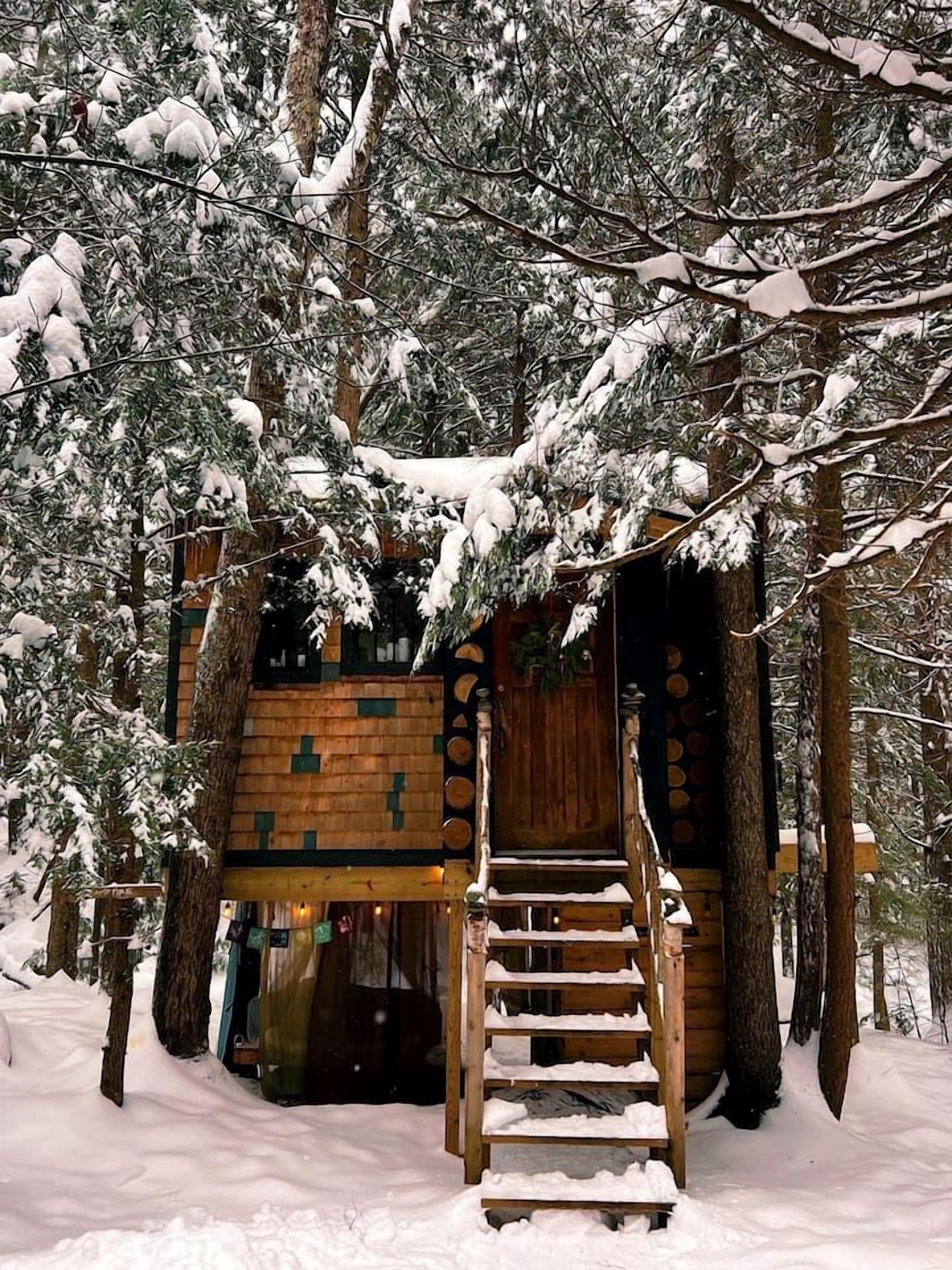 Eine Holzhütte mit einer Treppe, die zu einer Tür führt, umgeben von schneebedeckten Bäumen.