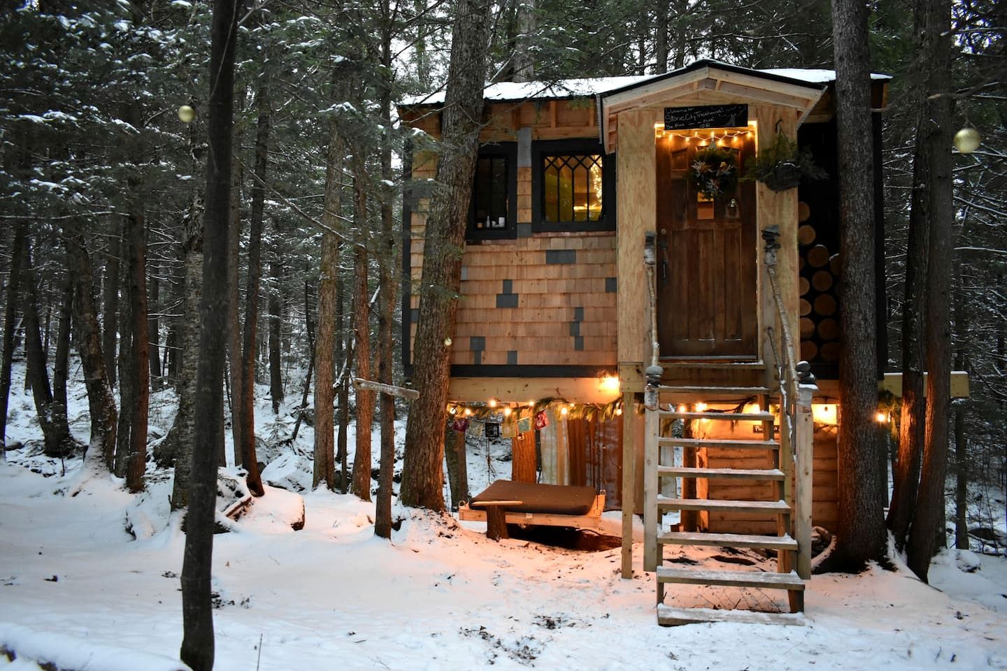 A wooden cabin with a staircase leading to a deck, surrounded by a snowy forest.