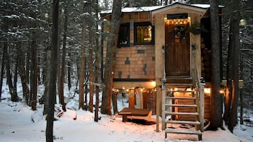 A wooden cabin with a staircase leading to a deck, surrounded by a snowy forest.