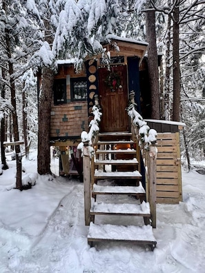 A wooden cabin with a staircase leading to a door, surrounded by snow-covered trees.