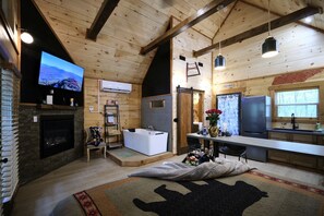 Interior de una cabaña de troncos con chimenea, televisor grande, bañera, zona de cocina y mesa de comedor.