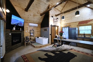 Ein rustikales Blockhaus-Interieur mit Kamin, großem Fernseher, Badewanne, Küchenbereich und Esstisch.