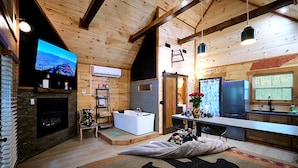 A log cabin interior with a stone fireplace, a large flat-screen TV, a wooden ceiling, a kitchen area, and a bathroom with a bathtub. - Stunning Cabin Nestled Amidst Trees with Hot-tub in Sevierville, Tennessee (Sevierville)