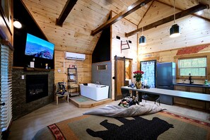 A log cabin interior with a stone fireplace, a large flat-screen TV, a wooden ceiling, a kitchen area, and a bathroom with a bathtub. - Stunning Cabin Nestled Amidst Trees with Hot-tub in Sevierville, Tennessee (Sevierville)