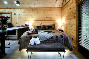 Ein Zimmer in einer Holzhütte mit einem Bett, einer Bank, einem Nachttisch und einem Fenster mit Jalousien.