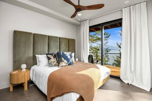 Une chambre à coucher comprenant un lit, une table de chevet, un ventilateur de plafond et une grande fenêtre avec des rideaux.