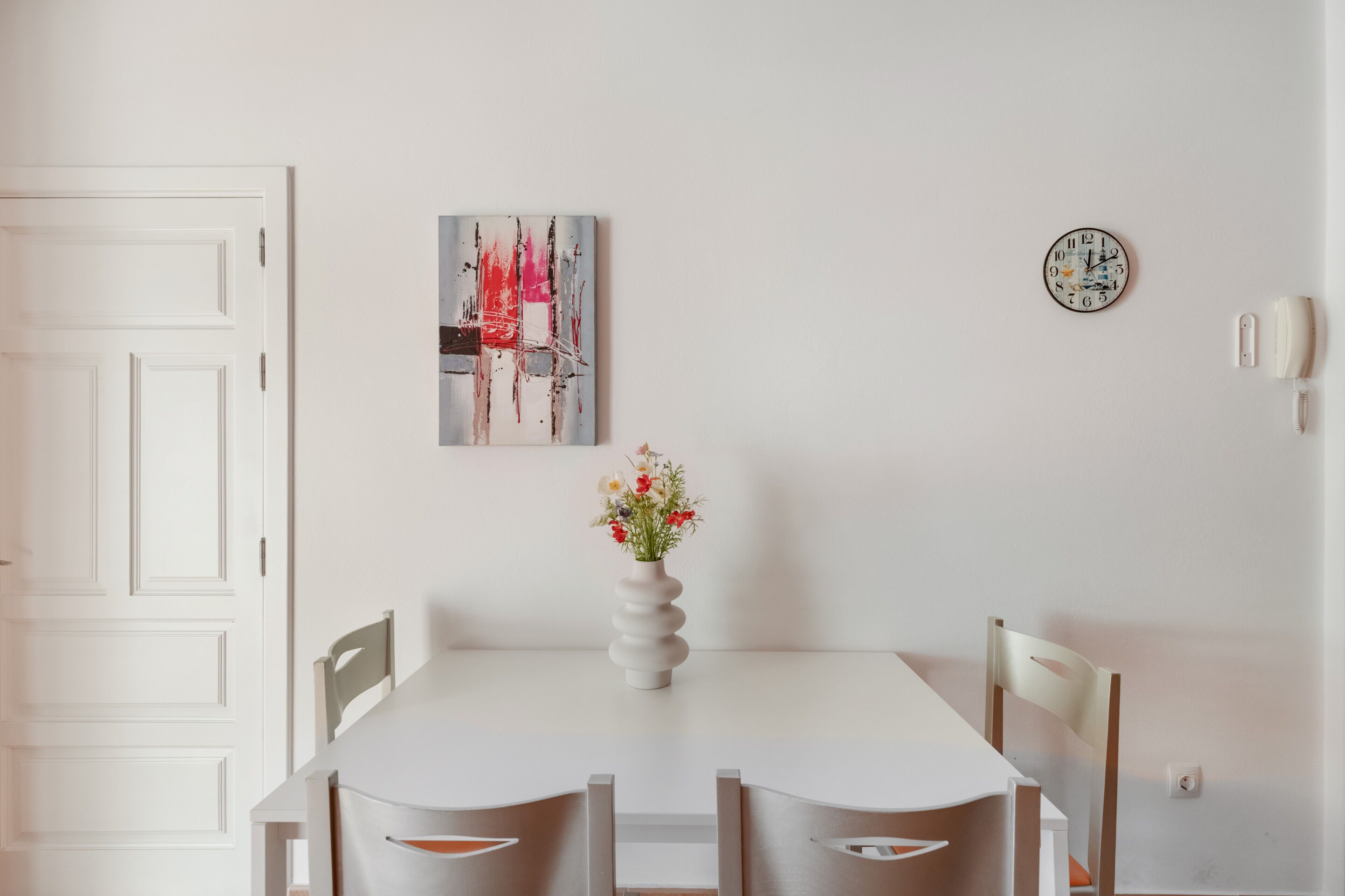 A dining area with a white table, two chairs, a vase with flowers, a clock, and a painting on the wall.