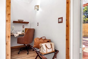 A room with a wooden desk, a brown leather chair, a leather bag on a small table, a wall-mounted light fixture, and a window with a view of greenery. - Impressive Intimate Tiny Cabin with Lofted Interior and Outdoor Shower in Texas (Wimberley)