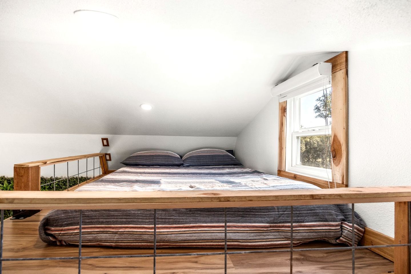 A bedroom with a sloped ceiling, a window with a view, a bed with striped bedding, and a wooden railing.